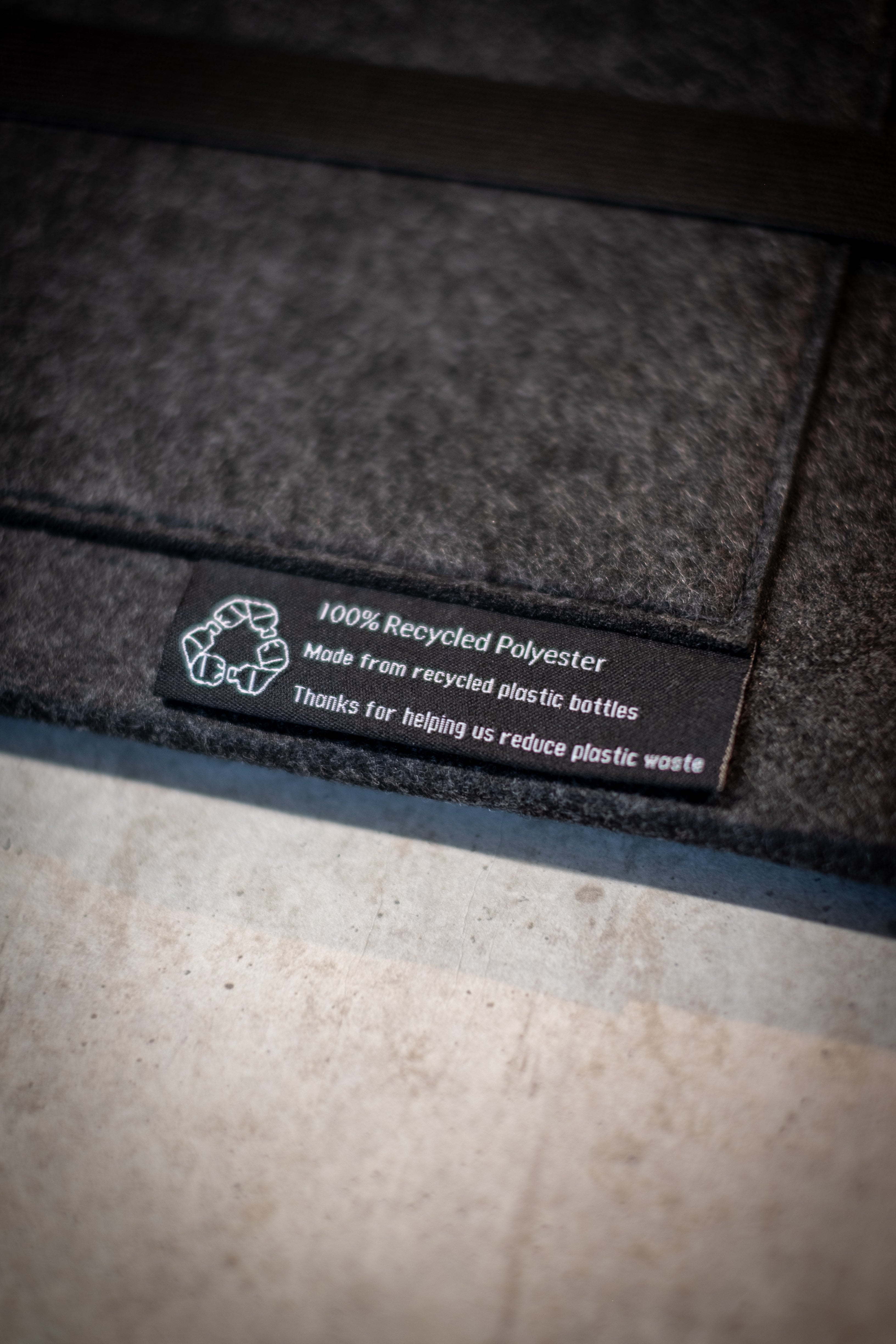 Close-up of the RMIT Store rPET Utility Laptop Sleeve showing the stitched black sustainability label that reads ‘100% Recycled Polyester – Made from recycled plastic bottles – Thanks for helping us reduce plastic waste’, against a charcoal felt background.